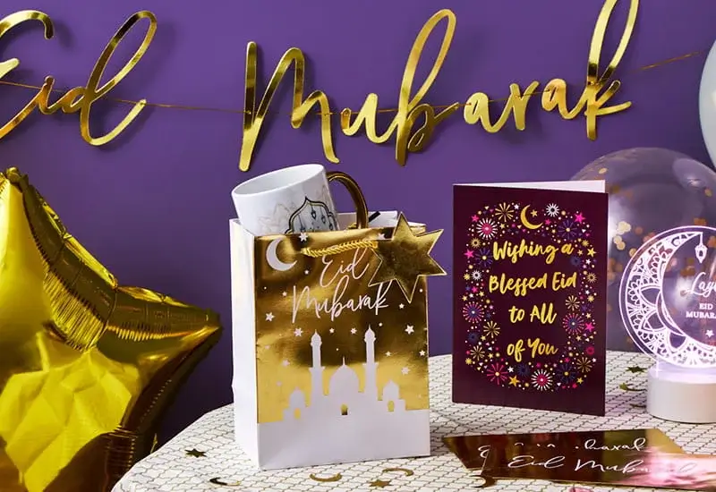 Gold and purple Eid-themed display featuring a gift bag with a mosque design, a mug, a greeting card with festive patterns, a star-shaped balloon, and Eid Mubarak decorations in the background.